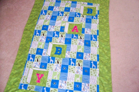 Baby Blanket, size is 38" x 54" embroidered applique for a crib cover, stroller blanket, made of all felt. The letters BABY are appliqued in Blue and Pink and embroidered as a block, on material design of numbers and giraffes in squares of white, yellow and blue with a green border. Backing of blue with poke a dots - Borgmanns Creations - 1