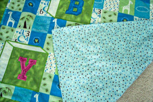 Baby Blanket, size is 38" x 54" embroidered applique for a crib cover, stroller blanket, made of all felt. The letters BABY are appliqued in Blue and Pink and embroidered as a block, on material design of numbers and giraffes in squares of white, yellow and blue with a green border. Backing of blue with poke a dots - Borgmanns Creations 