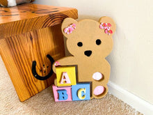 Load image into Gallery viewer, Baby shower gift - wood wall art teddy bear - wall hanging boy/ girl nursery decor - Shelf sitter or leaning against a wall at floor level. - this woodland nursery theme teddy bear is textured painted - pink flowers ion the ears and pink feet bottoms - pompom eyes and nose, - colorful ABC blocks - hanging hook on back - Borgmanns Creations
